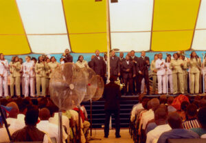Large group of people singing under a large yellow and white pole tent